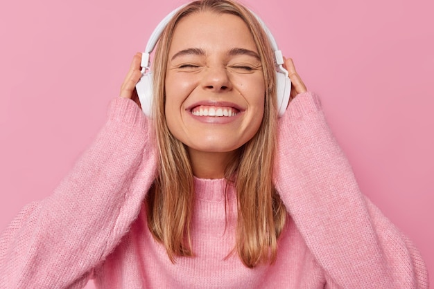 Garota do milênio feliz fecha os olhos e sorri para a câmera ouve música favorita através de fones de ouvido estéreo usa jumper macio confortável isolado sobre fundo rosa Pessoas estilo de vida e conceito de hobby