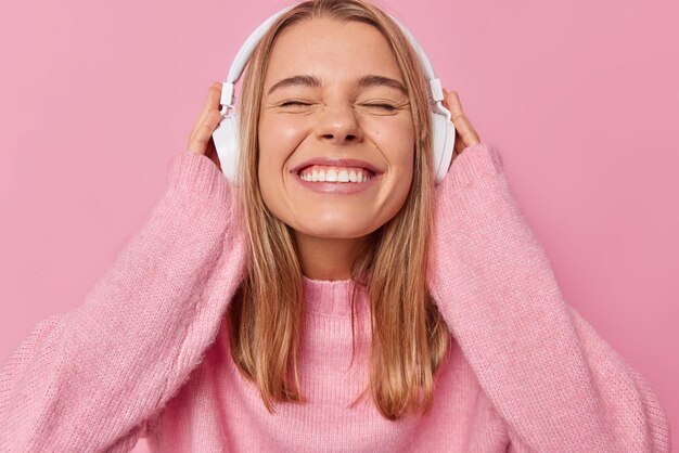 Garota do milênio feliz fecha os olhos e sorri para a câmera ouve música favorita através de fones de ouvido estéreo usa jumper macio confortável isolado sobre fundo rosa Pessoas estilo de vida e conceito de hobby