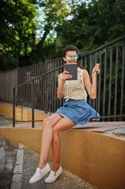 Garota elegante encaracolada usa blusa de saia jeans azul com tablet nas mãos