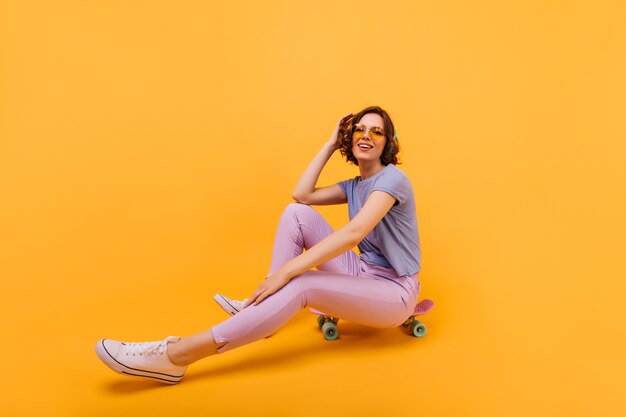 Garota em êxtase em calças rosa expressando boas emoções com longboard. Foto interna de atraente modelo feminino posando no skate.