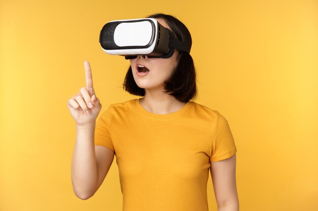 Garota em VR Linda jovem asiática usando óculos de realidade virtual e jogando conversando virtualmente sobre fundo amarelo