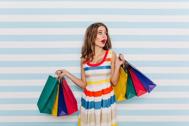 Garota emocional e surpresa em vestido listrado multicolorido, posando com sacolas de compras. Retrato de morena com batom vermelho na parede listrada