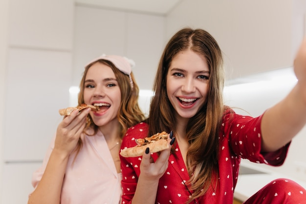 Foto grátis garota emocional encaracolada comendo pizza com um sorriso. foto interna de feliz mulher de cabelos castanhos em pijamas vermelha, fazendo selfie com um amigo.