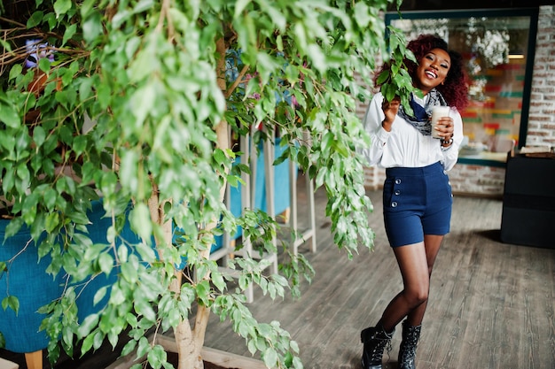 Garota encaracolada afro-americana atraente em blusa branca e shorts azuis posou no café contra árvore com café com leite à mão