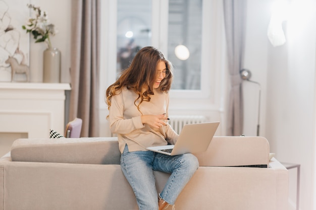 Garota encaracolada interessada usando laptop, sentada no sofá e rindo