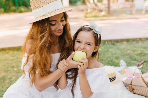 Garota engraçada dá uma mordida na grande maçã verde que está segurando sua linda mãe. Retrato ao ar livre de mulher jovem sorridente no elegante chapéu alimentando a filha com frutas saborosas em dia ensolarado.