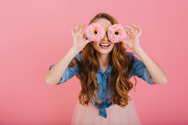 Garota engraçada e elegante em roupa da moda brinca com deliciosos donuts que comprou na padaria para o chá. Retrato de uma jovem graciosa e encaracolada posando com doces isolados em um fundo rosa