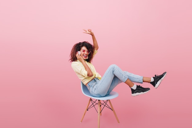 Garota engraçada em tênis preto e meias brancas, posando com interior rosa e ouvindo a música favorita. encantadora mulher africana em jeans da moda, sentada na cadeira.