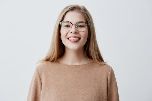 Garota engraçada otimista com cabelo liso loiro, vestindo blusa marrom e óculos, mostrando a língua enquanto posava contra fundo cinza studio. Emocional positivo jovem fêmea fazendo caretas