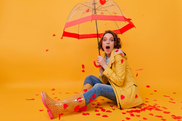 Garota entusiasmada surpresa com sapatos de outono de borracha, sentada no chão. Retrato de estúdio de feliz modelo feminino posando, rodeado por corações de papel.