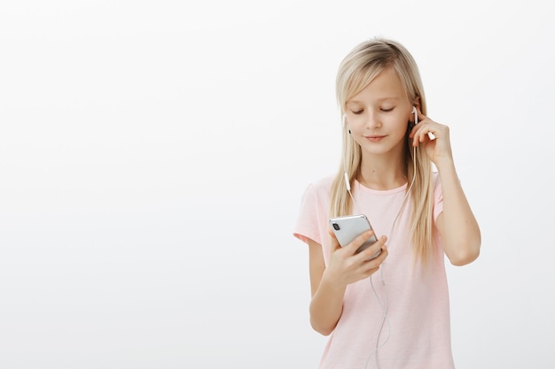 Garota esperta sabe tudo sobre gadgets. Retrato de uma linda criança loira linda, usando fones de ouvido e escolhendo música no smartphone