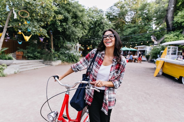 Garota espetacular com roupa casual, aproveitando o lazer ativo na primavera. modelo latino feminino em copos de pé na rua com a bicicleta.
