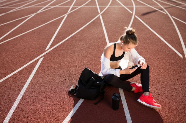 Garota esportiva em fones de ouvido sem fio com mochila e garrafa esportiva perto alegremente usando o celular enquanto passa o tempo na esteira do estádio