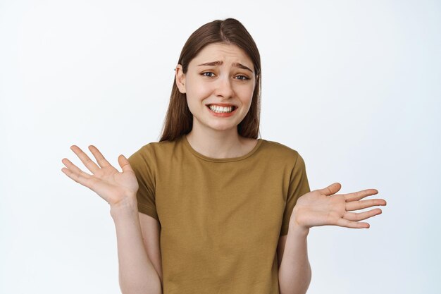 Garota estranha encolhendo os ombros com expressão de rosto se desculpando, parecendo sem noção, peça desculpas, mostrando as mãos vazias, não tem nada, de pé sobre fundo branco.