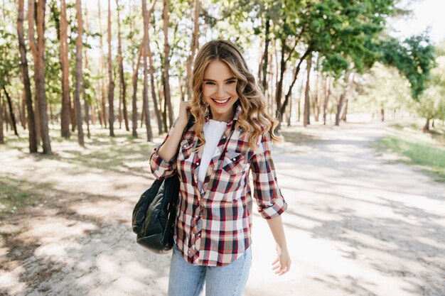 Garota europeia confiante andando no parque com a mochila. Tiro ao ar livre de romântica senhora loira usa camisa quadriculada na primavera.