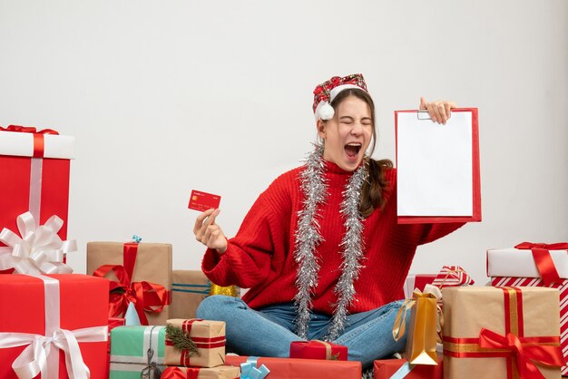 Foto grátis garota exultante com chapéu de papai noel segurando um cartão e um documento sentado em frente aos presentes