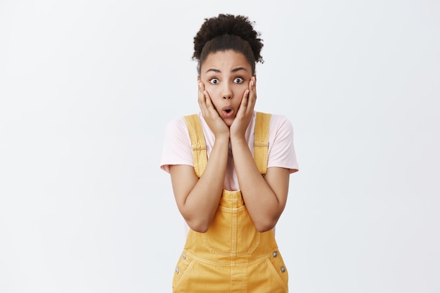 Foto grátis garota fácil de surpreender com rumores chocantes. retrato de uma aluna afro-americana impressionada, espantada e surpreendida de macacão amarelo com cabelo encaracolado penteado, ofegando por tremer e olhando espantada