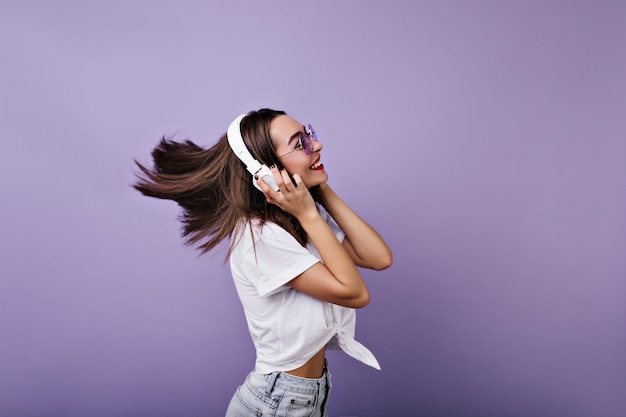 Garota fascinante com cabelo liso e brilhante dançando e rindo. Retrato de uma jovem engraçada em fones de ouvido isolados.