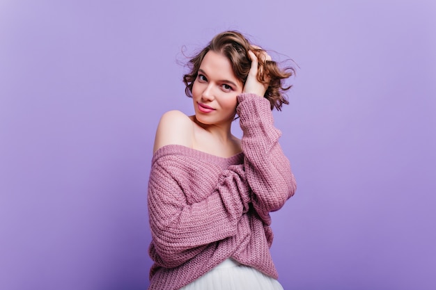 Garota fascinante em suéter de lã brinca com cabelo curto brilhante. elegante mulher branca com penteado da moda isolado na parede roxa.