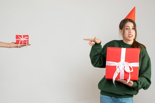 garota feliz com boné de festa segurando seu presente de natal apontando para uma mão humana segurando um presente em branco