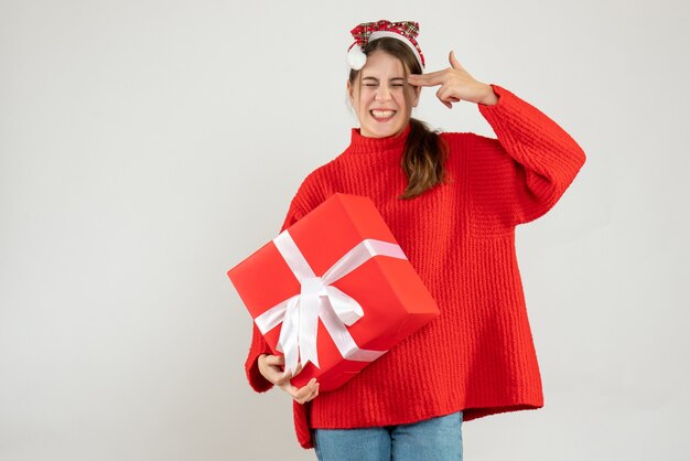 garota feliz com chapéu de Papai Noel segurando um presente e colocando a arma de dedo na têmpora branca