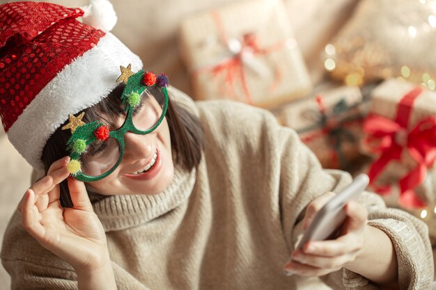 Garota feliz de óculos engraçados de Natal e chapéu de Papai Noel com um telefone nas mãos.