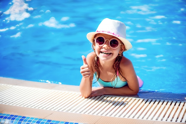 Garota feliz em óculos de sol e chapéu com unicórnio aparecer o polegar na piscina do resort de luxo nas férias de verão na ilha de praia tropical