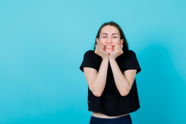 Garota feliz está sorrindo segurando os punhos sob o queixo no fundo azul