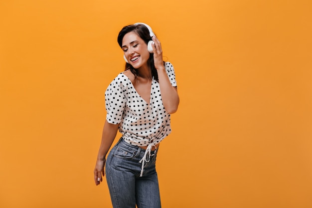 Foto grátis garota feliz na blusa de bolinhas ouve música com fones de ouvido em fundo laranja. menina adulta sorridente em jeans e camisa leve gosta.