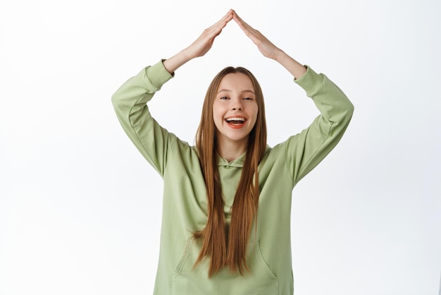 Garota feliz sorridente mostra gesto de telhado em casa com as mãos acima da cabeça rindo e parecendo otimista fazendo sinal de telhado em pé com capuz contra fundo branco