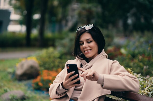 Garota feliz, usando um telefone inteligente em um parque da cidade, sentado num banco