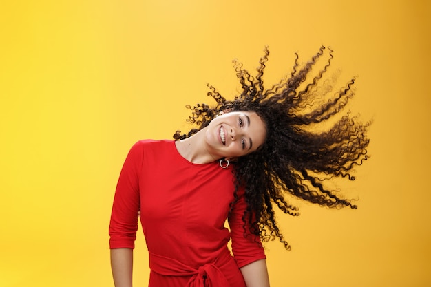 Garota ficando despreocupada e selvagem balançando o cabelo e fazendo sol com cachos voando no ar sorrindo amplamente inclinando a cabeça e se divertindo, dançando encantada e divertida, sentindo-se lúdica contra o fundo amarelo.