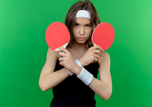 Garota fitness jovem em roupa esportiva preta com tiara segurando duas raquetes de tênis de mesa descontente cruzando as mãos em pé sobre a parede verde