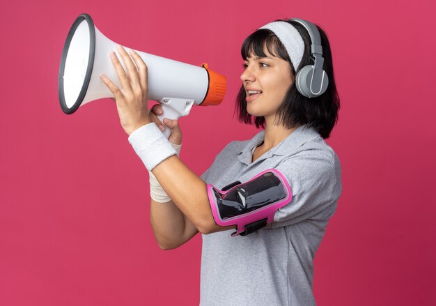 Garota fitness jovem usando bandana com fones de ouvido e uma braçadeira para smartphone gritando para o megafone feliz e confiante em pé sobre um fundo rosa