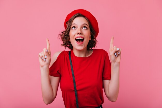 Garota francesa em êxtase com tatuagens sorrindo. Mulher elegante surpresa na boina vermelha, olhando para cima.