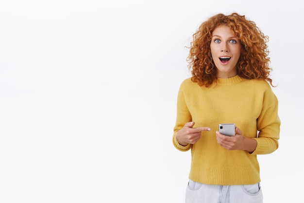 Garota gengibre com cabelo encaracolado natural, segurando um smartphone, apontando o telefone celular e olhando para frente impressionada