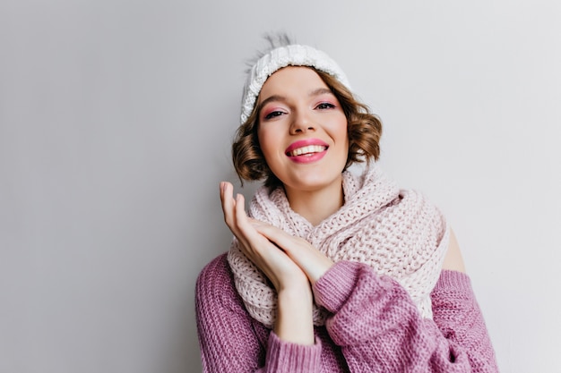 Garota glamourosa com maquiagem rosa, posando em acessórios de inverno de malha na parede de luz. Foto interna de uma mulher de cabelos curtos entusiasmada com chapéu e lenço.