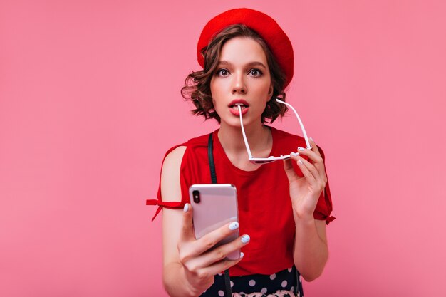 Garota graciosa em êxtase com óculos escuros posando de brincadeira. Mulher francesa encantadora com smartphone em pé na mão.