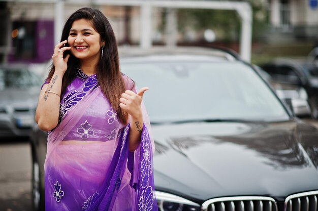 Garota hindu indiana no tradicional saree violeta posou na rua contra o carro suv de negócios preto e falando no celular mostra o polegar para cima