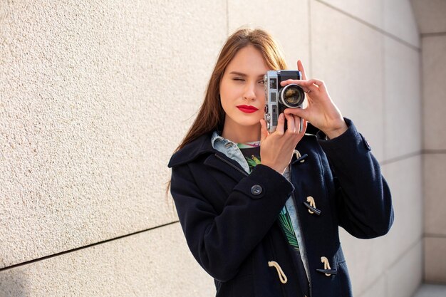 Garota hipster feliz fazendo foto com câmera retro na rua da cidade. Conceito de viagem