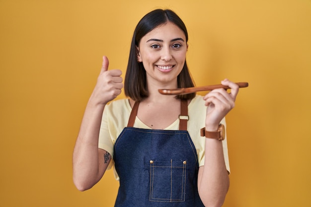 Garota hispânica comendo colher de pau saudável fazendo gesto feliz com polegares para cima com expressão de aprovação de mão olhando para a câmera mostrando sucesso