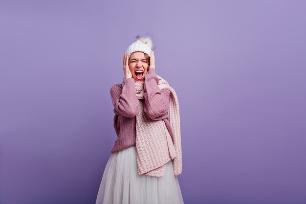 Garota incrível em cachecol de malha comprida gritando com os olhos fechados. Magnífica senhora europeia com roupas de inverno elegantes, posando na parede roxa