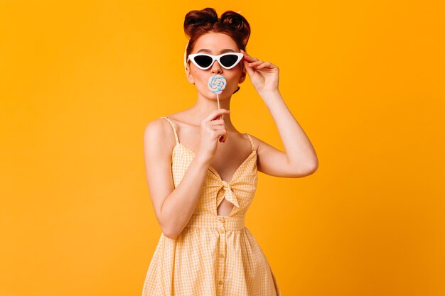 Garota incrível em óculos de sol, lambendo o pirulito. Foto de estúdio de gengibre pin-up mulher isolada no espaço amarelo.