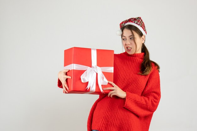 Garota insatisfeita com um chapéu de Papai Noel segurando um presente de pé