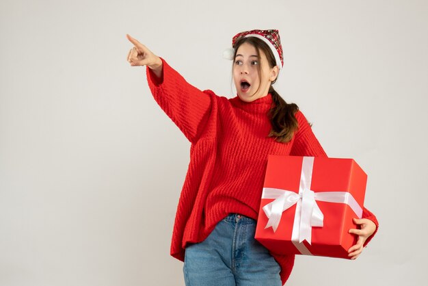 garota interessada com chapéu de Papai Noel segurando o dedo no presente e apontando algo em pé no branco