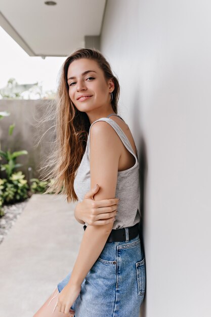 Garota interessada usa saia jeans e faixa preta em pé perto da parede branca. Mulher espetacular de cabelos compridos em traje casual, posando com expressão facial inspirada em bom dia.