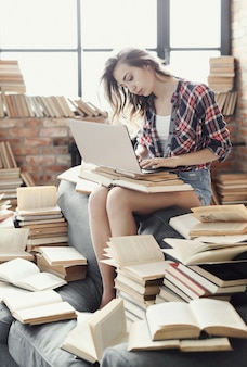 Garota jovem adolescente usando o computador portátil, rodeado por muitos livros.
