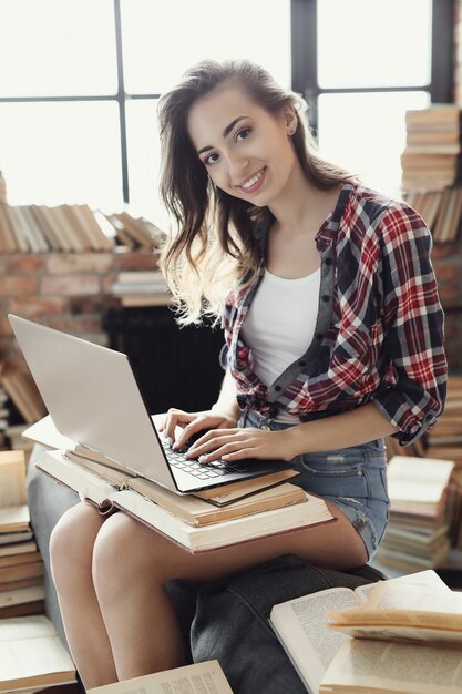 Garota jovem adolescente usando o computador portátil, rodeado por muitos livros.