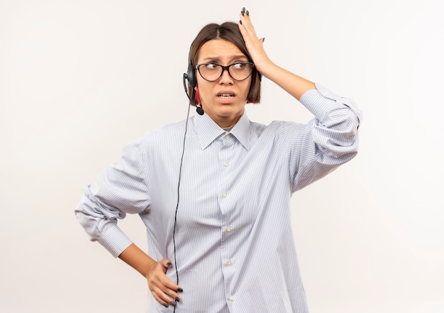 Garota jovem confusa de call center usando óculos e fone de ouvido colocando as mãos na cabeça e na cintura, olhando para o lado isolado no branco