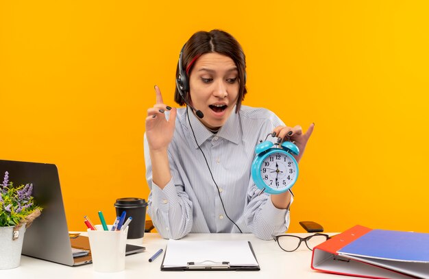 Garota jovem de call center impressionada usando fone de ouvido sentada na mesa, segurando e olhando para o despertador e levantando o dedo isolado em laranja